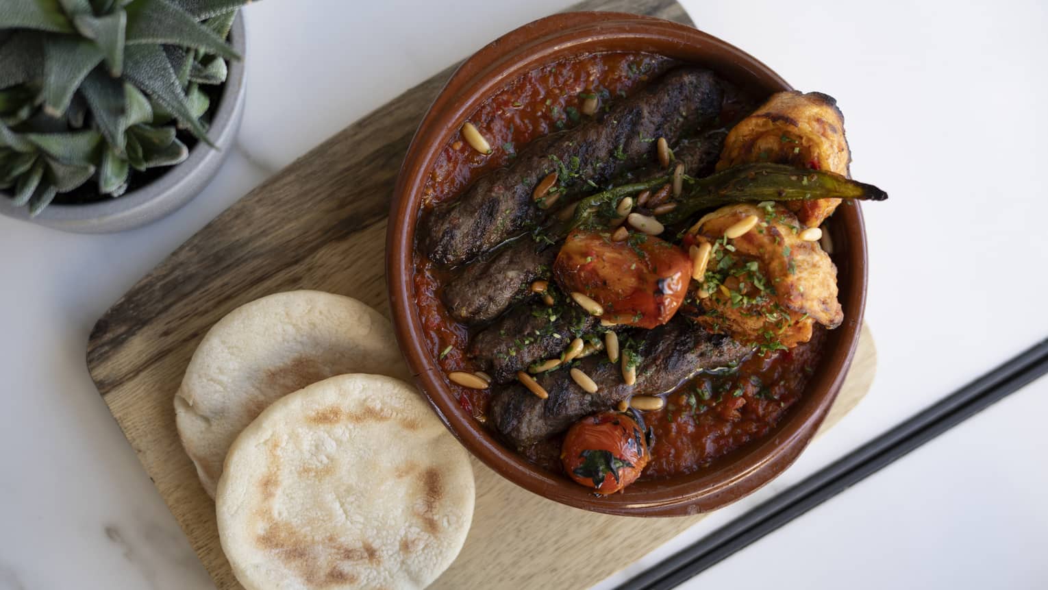 Kebab Khashkhash served in a brown stoneware bowl next to two small pieces of naan all on a wood serving tray