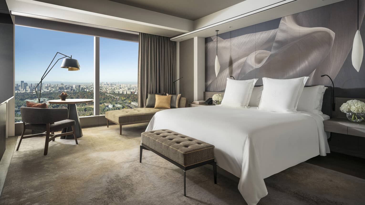 Guest room with king bed, tufted bench, chaise lounge, table with armchair, pendant lights and large window with city views