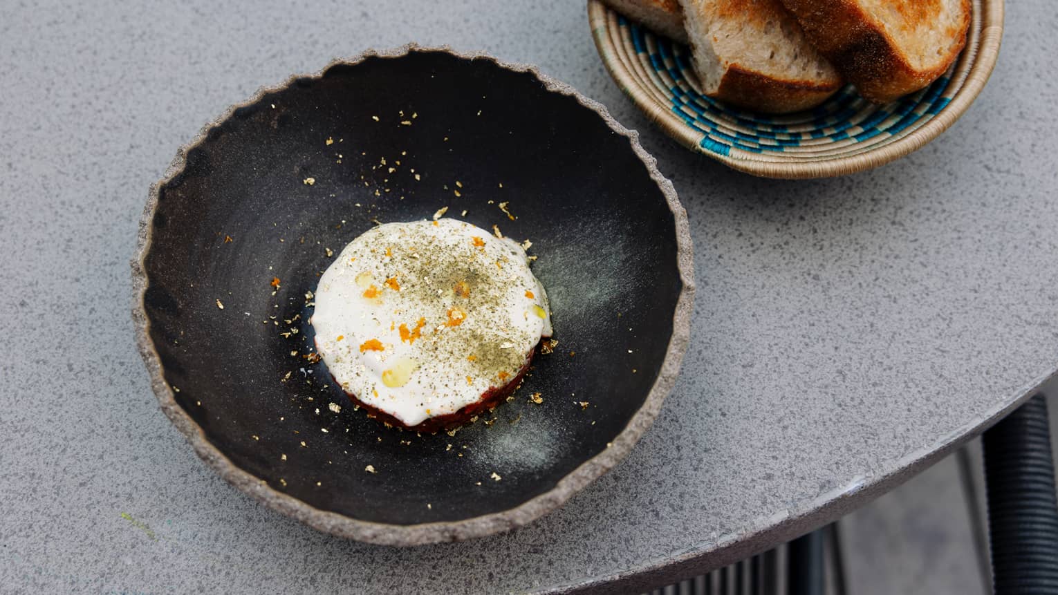An artistic black bowl holds food items. A plate of bread slices sits next to it.