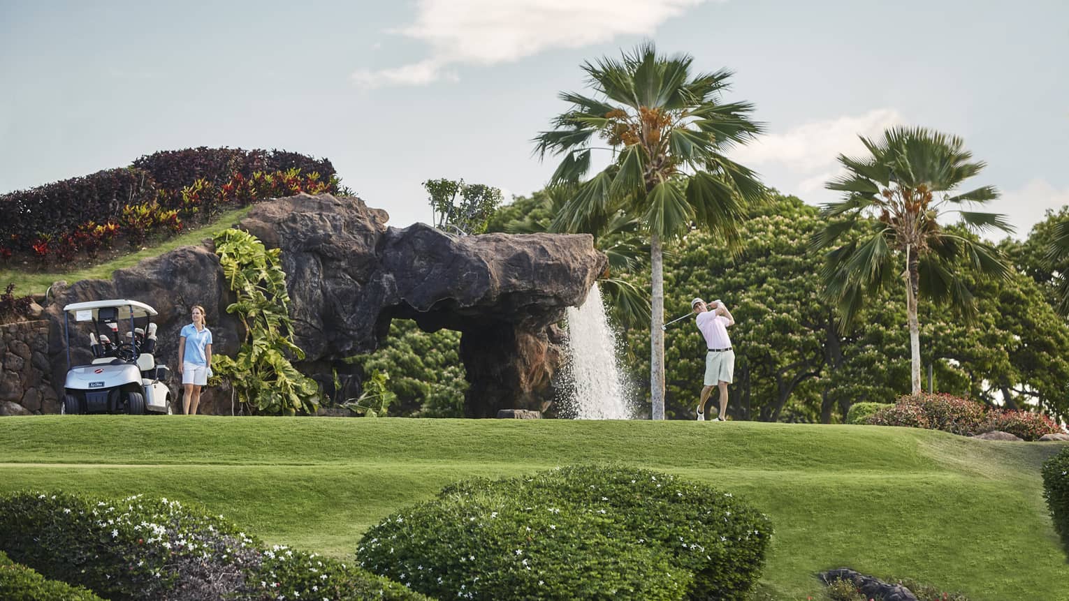Man swings golf club on course by rock and waterfall, woman stands by cart
