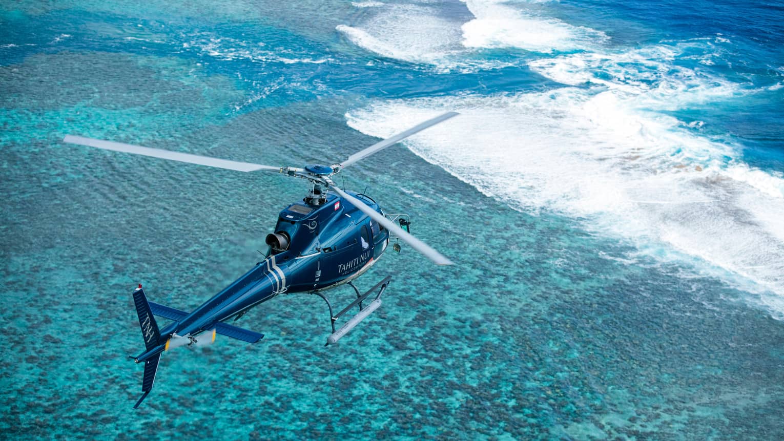 Bird’s-eye view of a helicopter flying over crystal-clear teal water with frothy waves breaking over a sandbar.