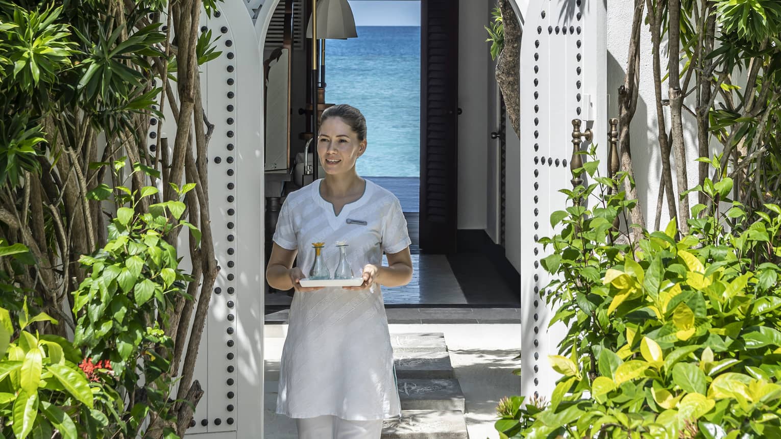 Therapist carrying a tray of oils through the spa entrance framed by an ornate archway and green foliage, with a view of the turquoise water in the background