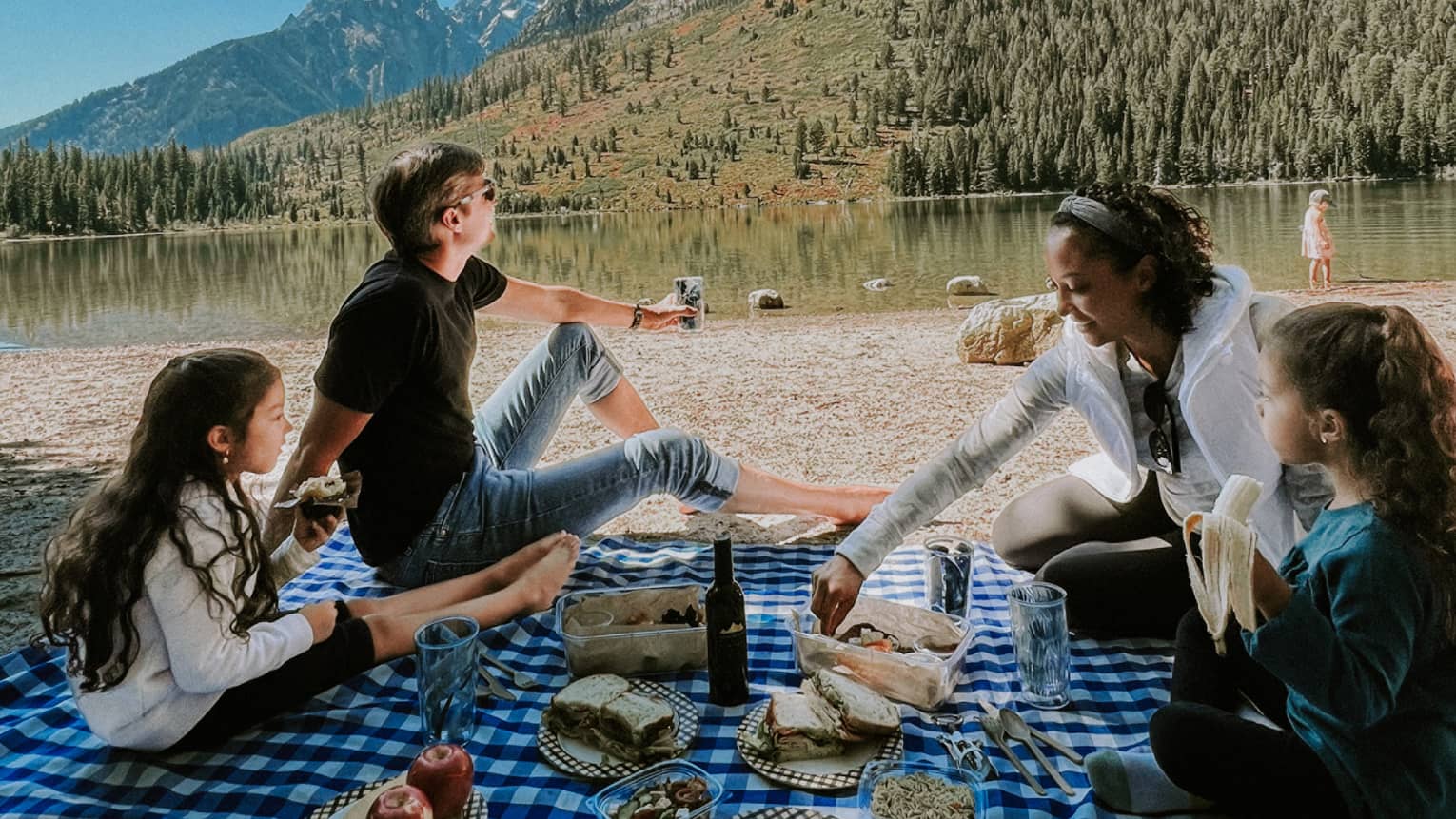 A family picnics on a checkered blanket near an alpine lake reflecting a rust and gold treed hill, distant mountains beyond,.