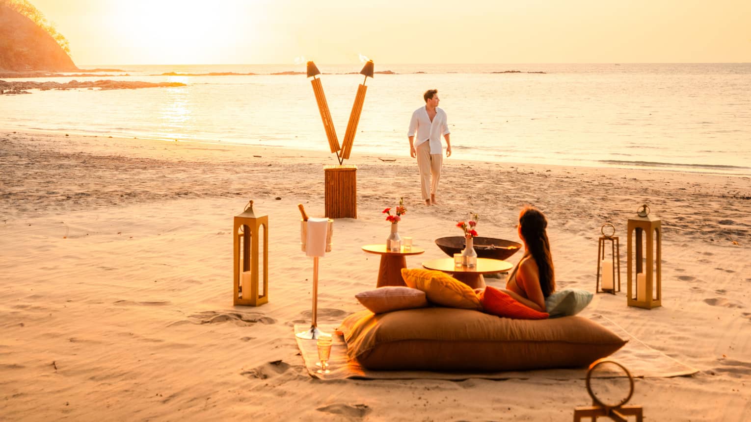 One person lounges on a large cushion set on a blanket on the sand while another walks back from the ocean to a private dining set up