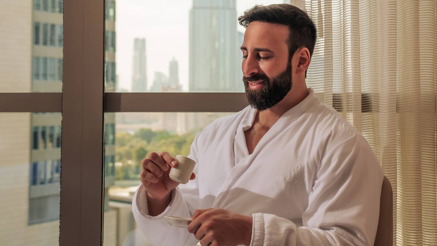 Person with short dark hair and beard and wearing a white spa bath robe sits by a window sipping tea with a city skyline in the background