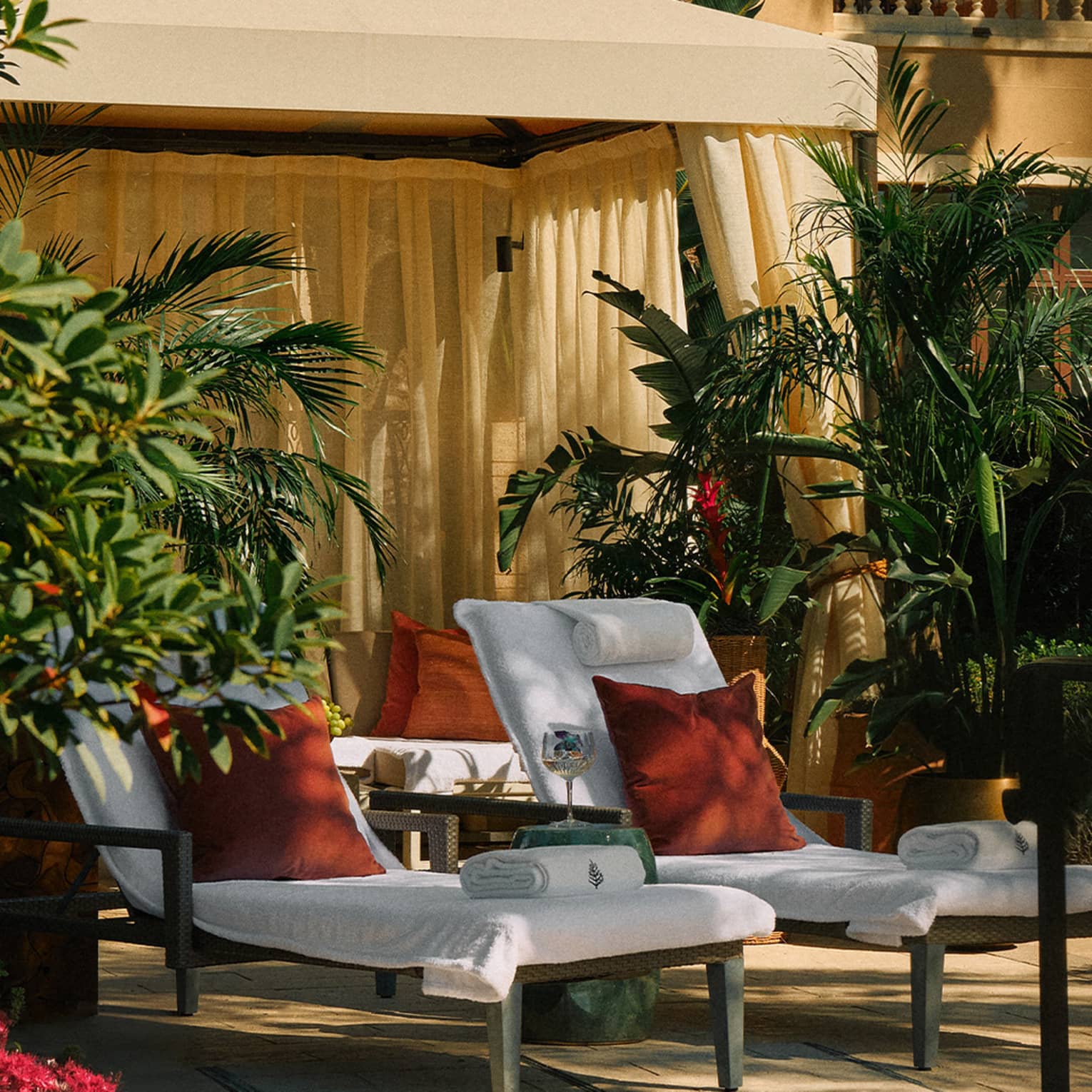 Two lounge chairs under a cabana by the pool surrounded by plants