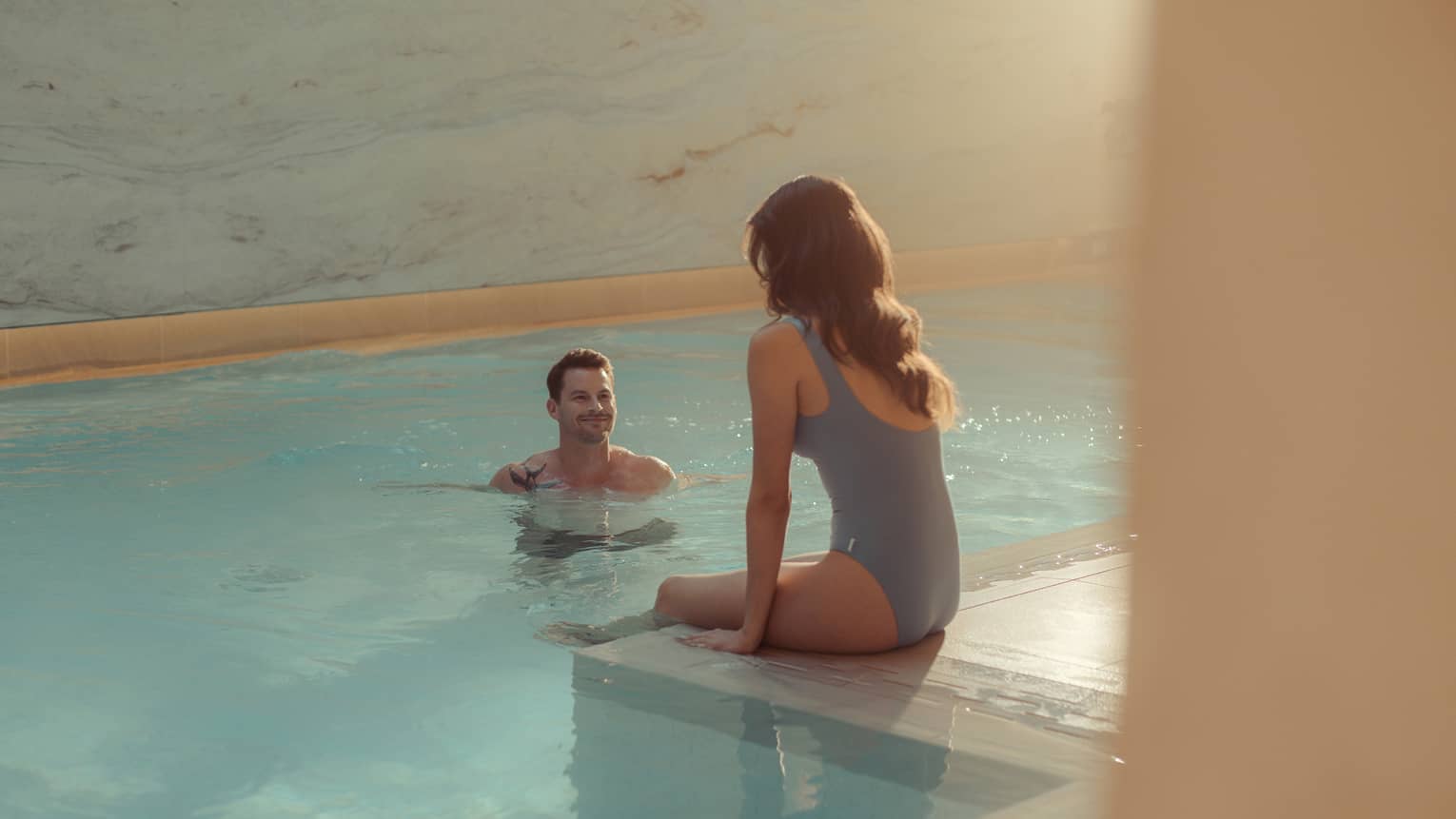 At an indoor pool with marble walls illuminated by natural sunlight, a woman sits on the pool's tiled edge as a man stands in the clear, blue water facing her