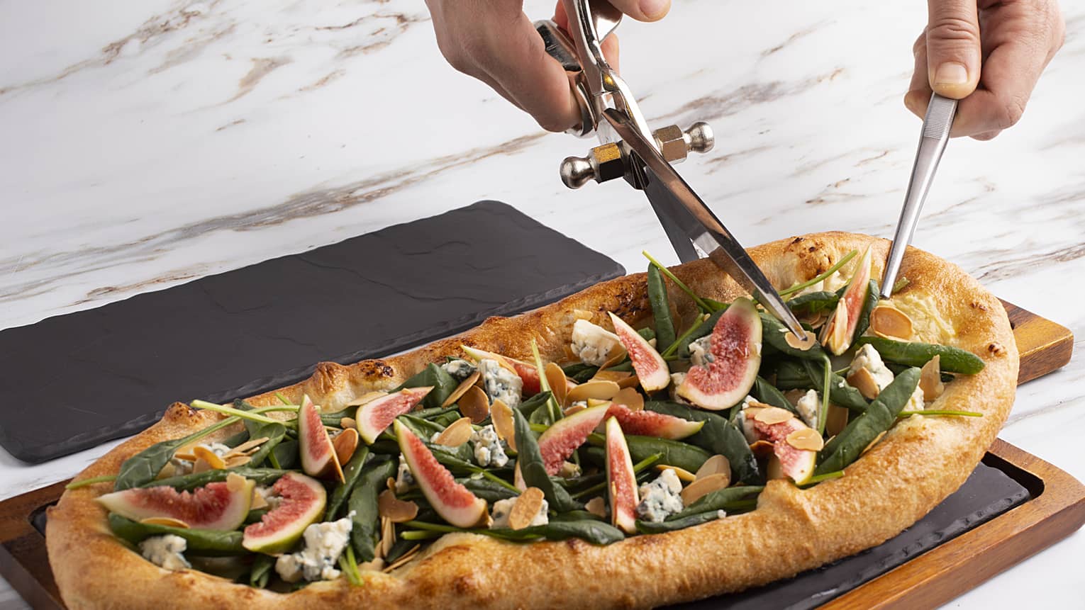 A large pizza topped with various fruits and vegetables, being cut into with a knife and pair of scissors.