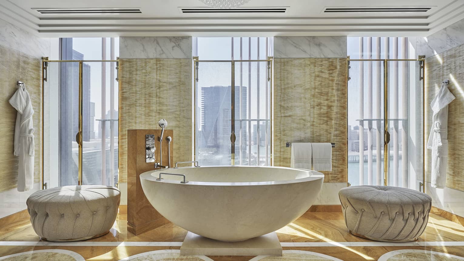 Luxurious bathroom with a freestanding stone bathtub, tufted ottomans, golden accents and large windows offering city and water views