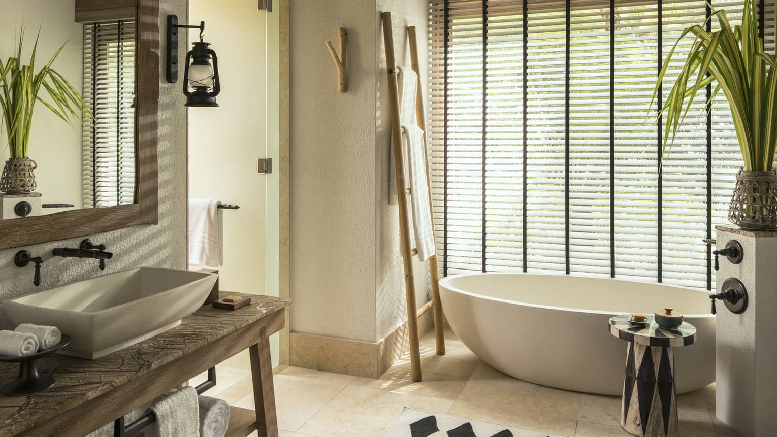 Free-standing white tub in bathroom under window with closed blinds, sink and vanity