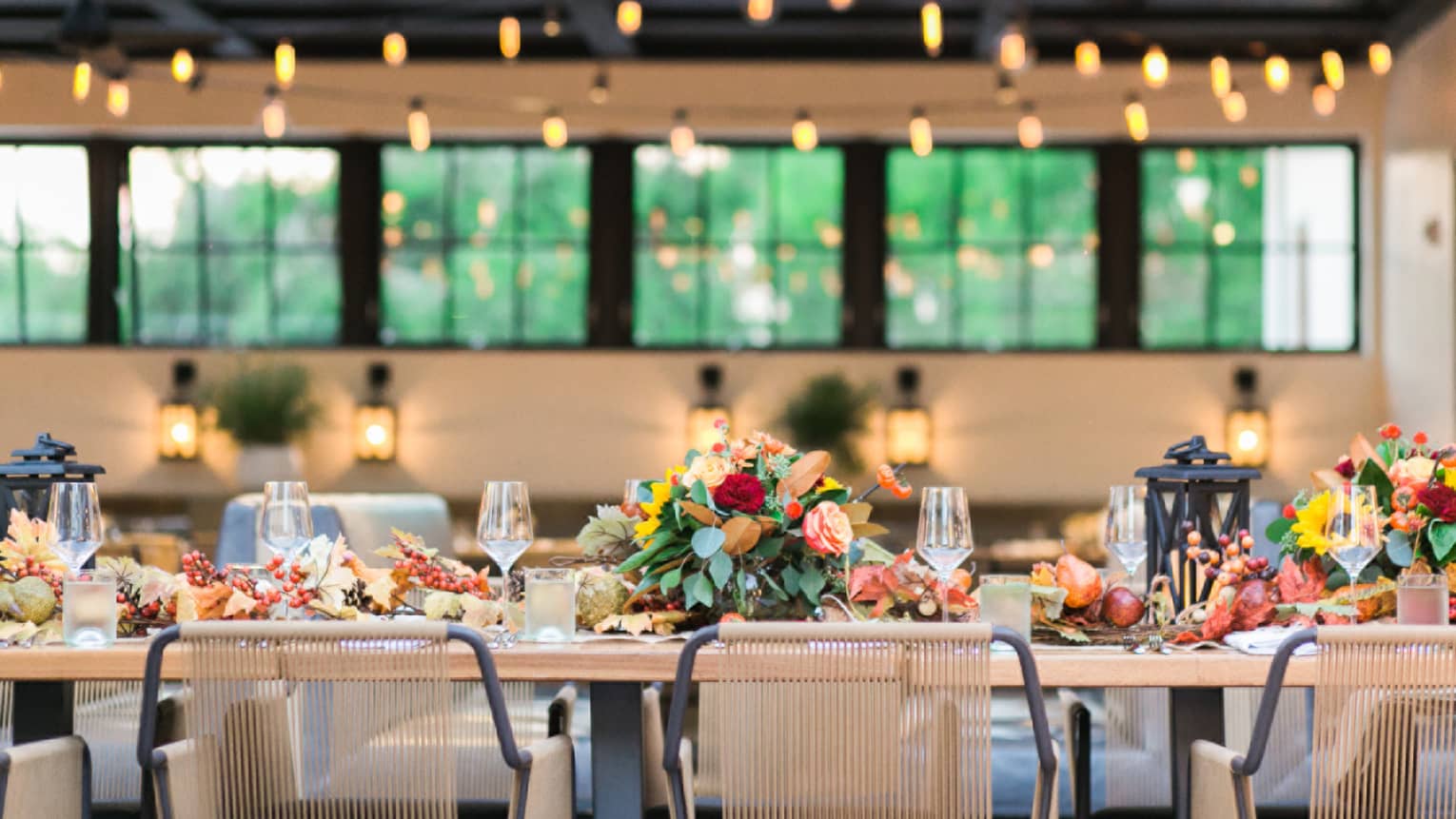 Long outdoor dining table with harvest flower arrangements under strings of bulb lights during day