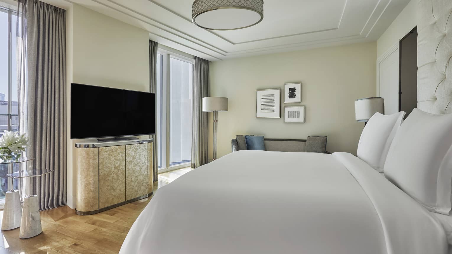 Bright hotel bedroom with a plush white bed, a flat-screen TV on a sleek cabinet, modern decor and large windows with gray curtains