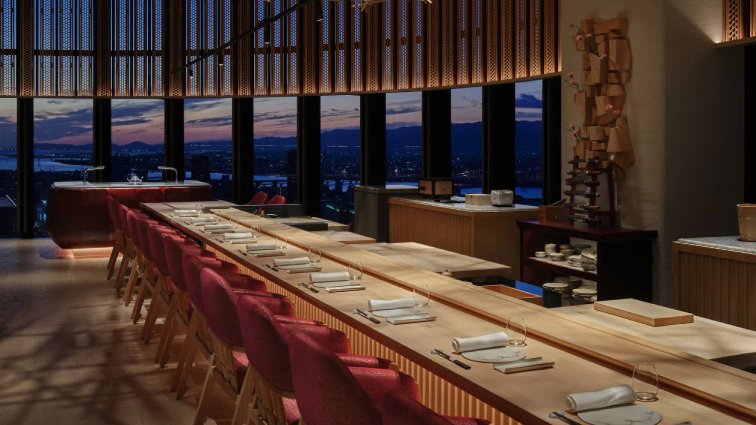 Ten place settings at a long wooden table with warm underlighting, a curved bank of windows offering a dramatic sunset view.