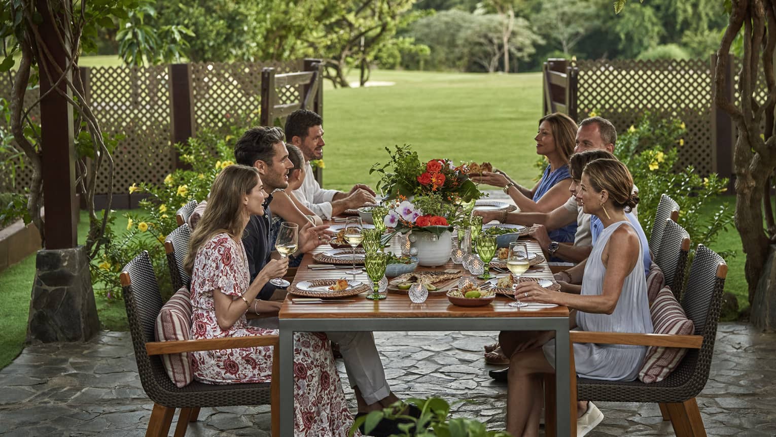 Eight people sit around a long dining table set up outside beneath a canopy of greenery and string lights