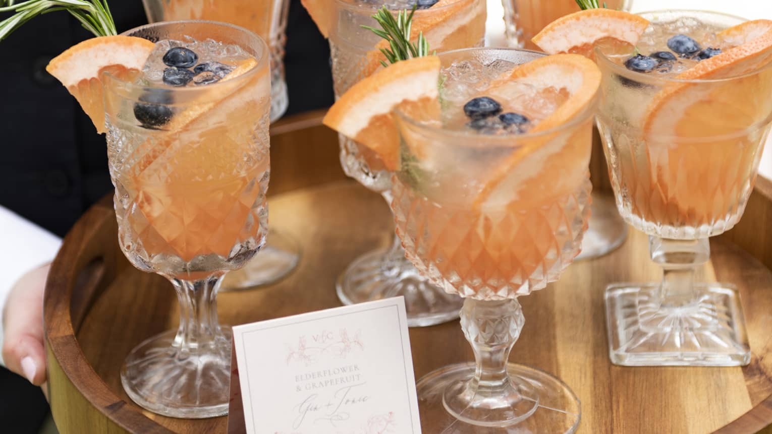 Hotel staff holds wood tray with six fruity cocktails in crystal glasses