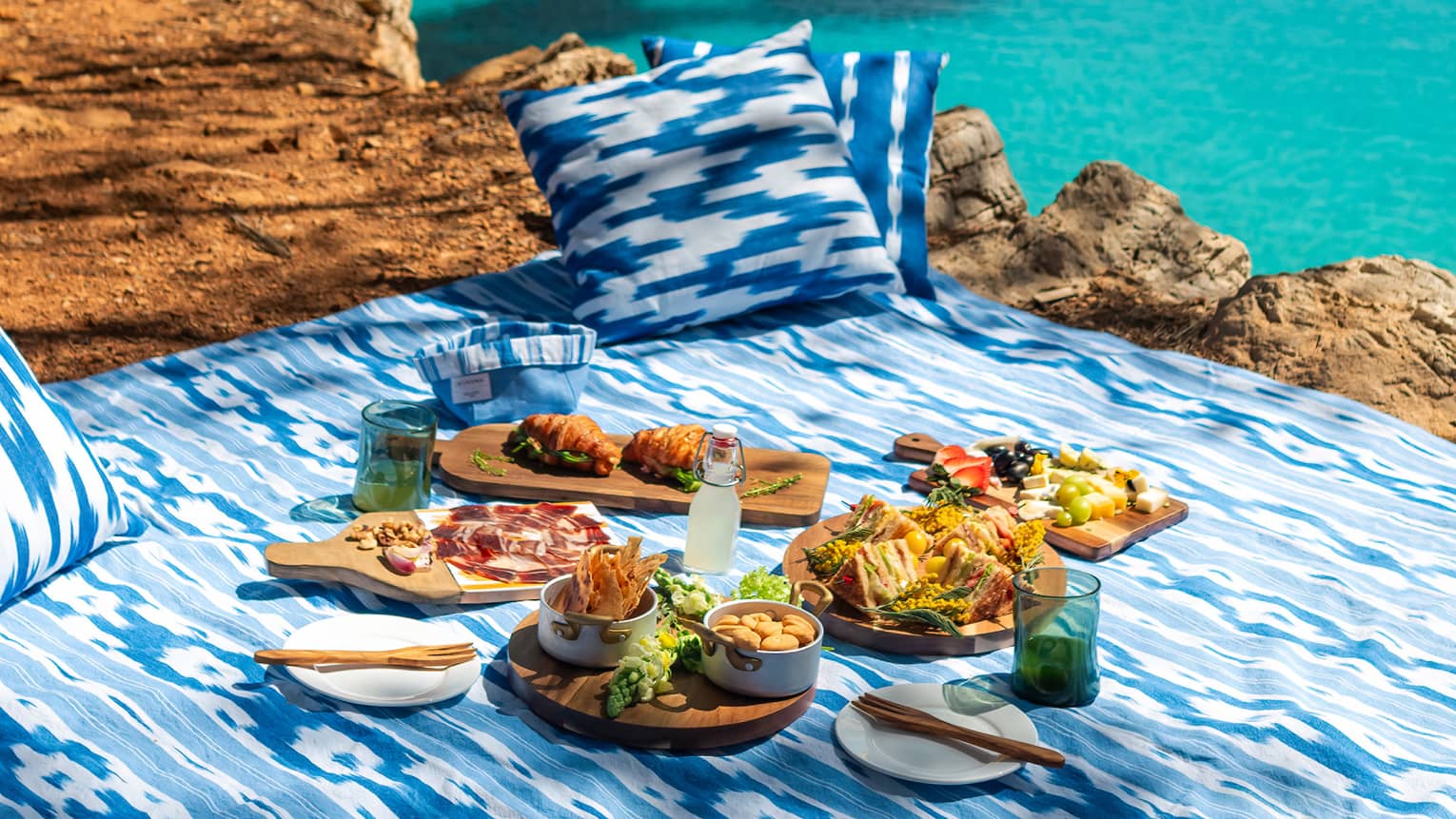 A picnic meal set out on a blue and white blanket with blue and white pillows, right next to the ocean. Turquoise waters, the rocky shoreline with trees, and blue sky are in the background.