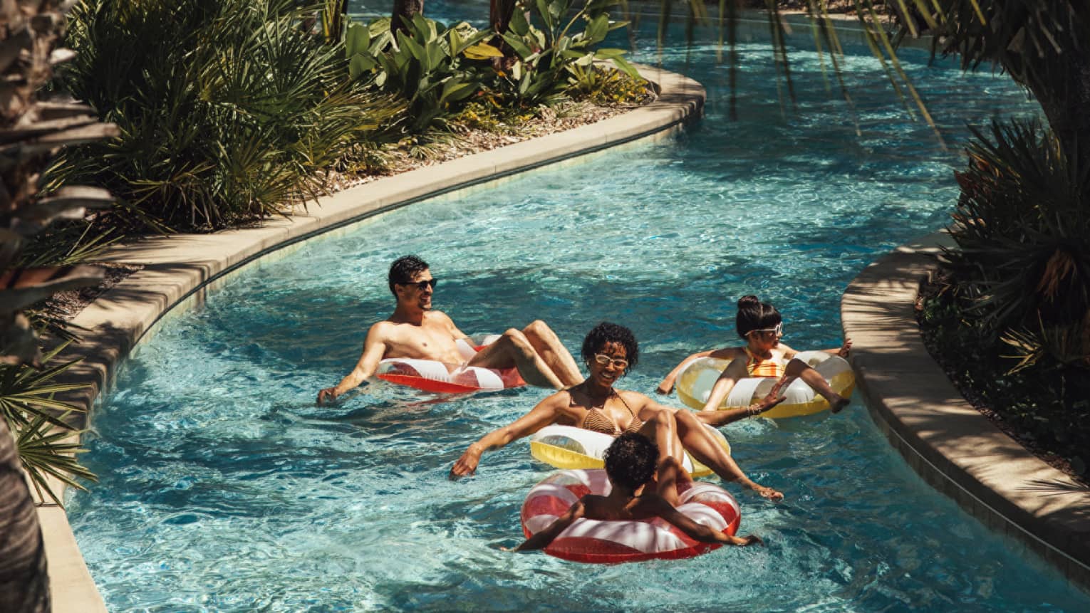 Two adults and two young children float in inner tubes down a lazy river surrounded by palm trees