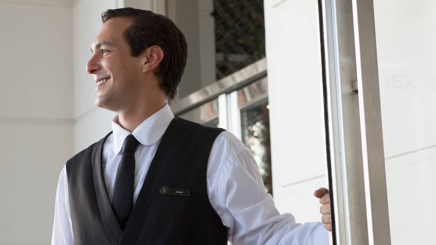 Man in black vest and tie, white shirt smiles and holds door open