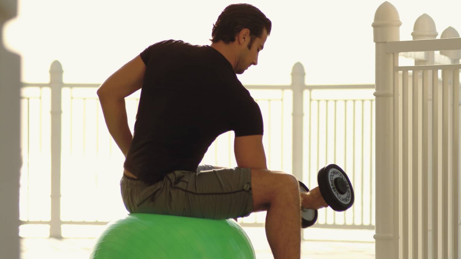 Back view of man in workout gear with hand weight, seated on exercise ball