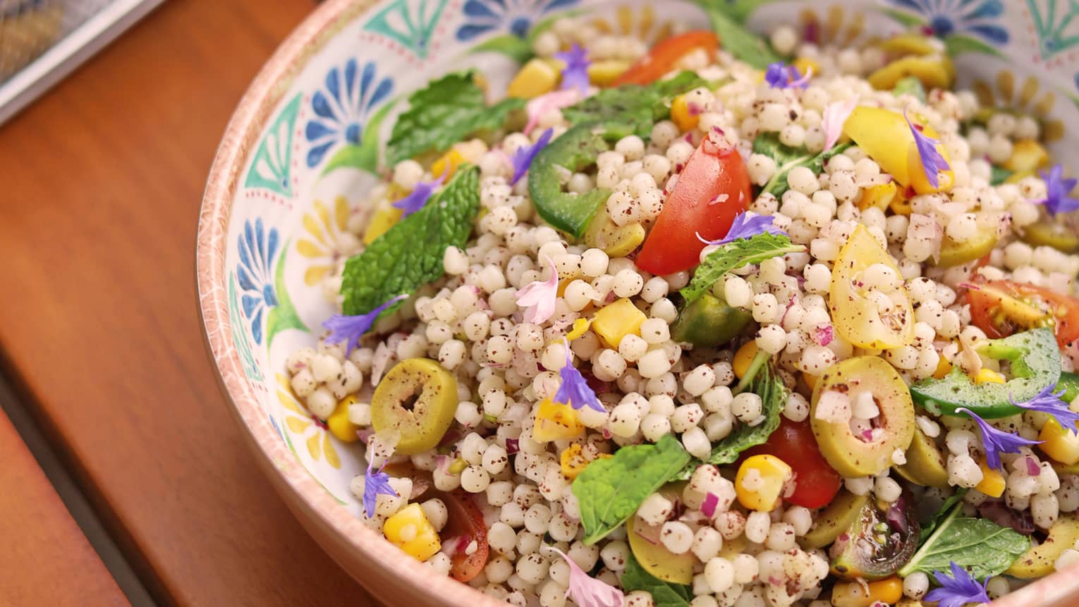 Couscous salad served in a patterend bowl next to a red-coloured beverage on a wooden table
