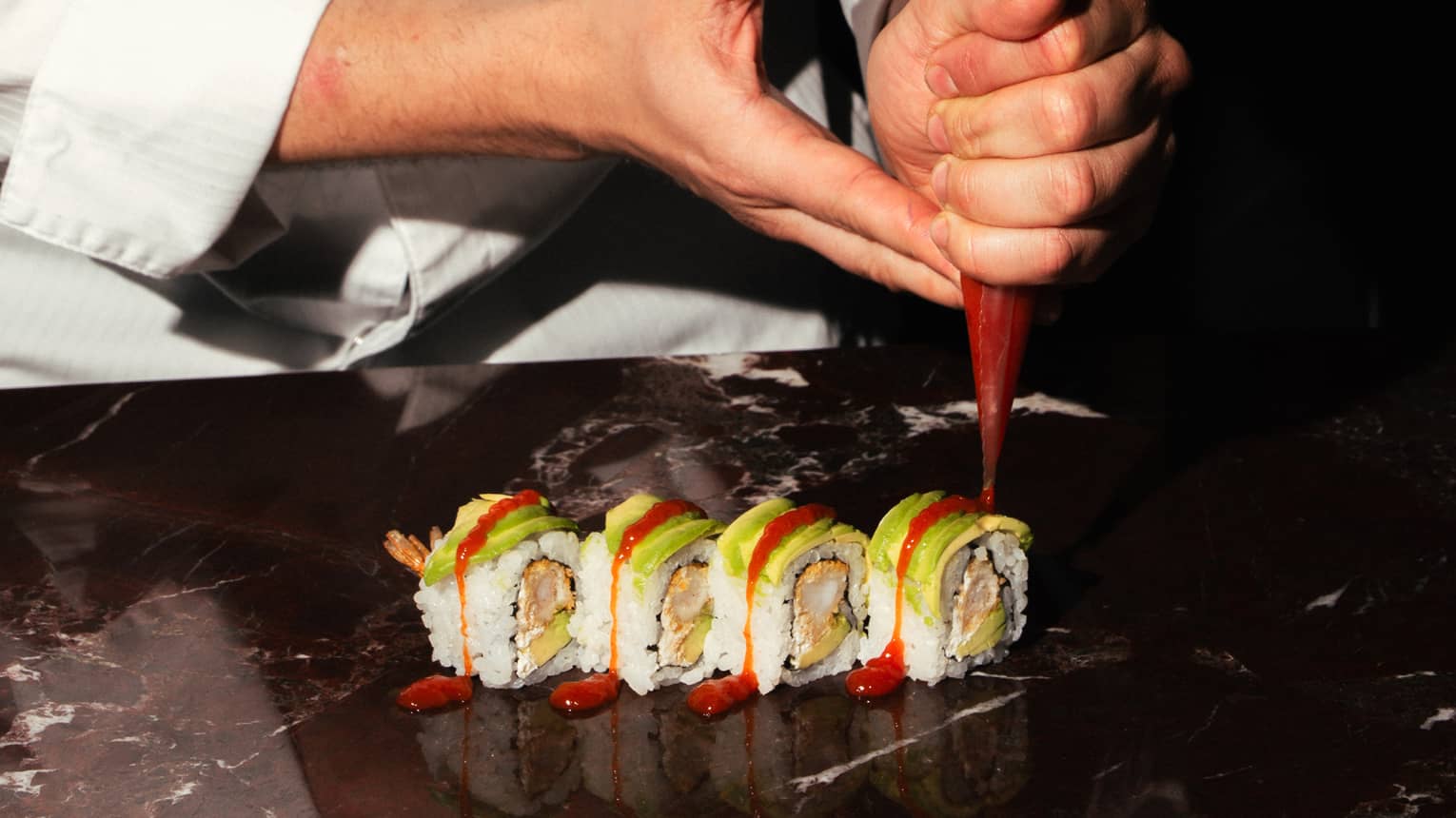 A chef adds garnish to a row of sushi rolls.