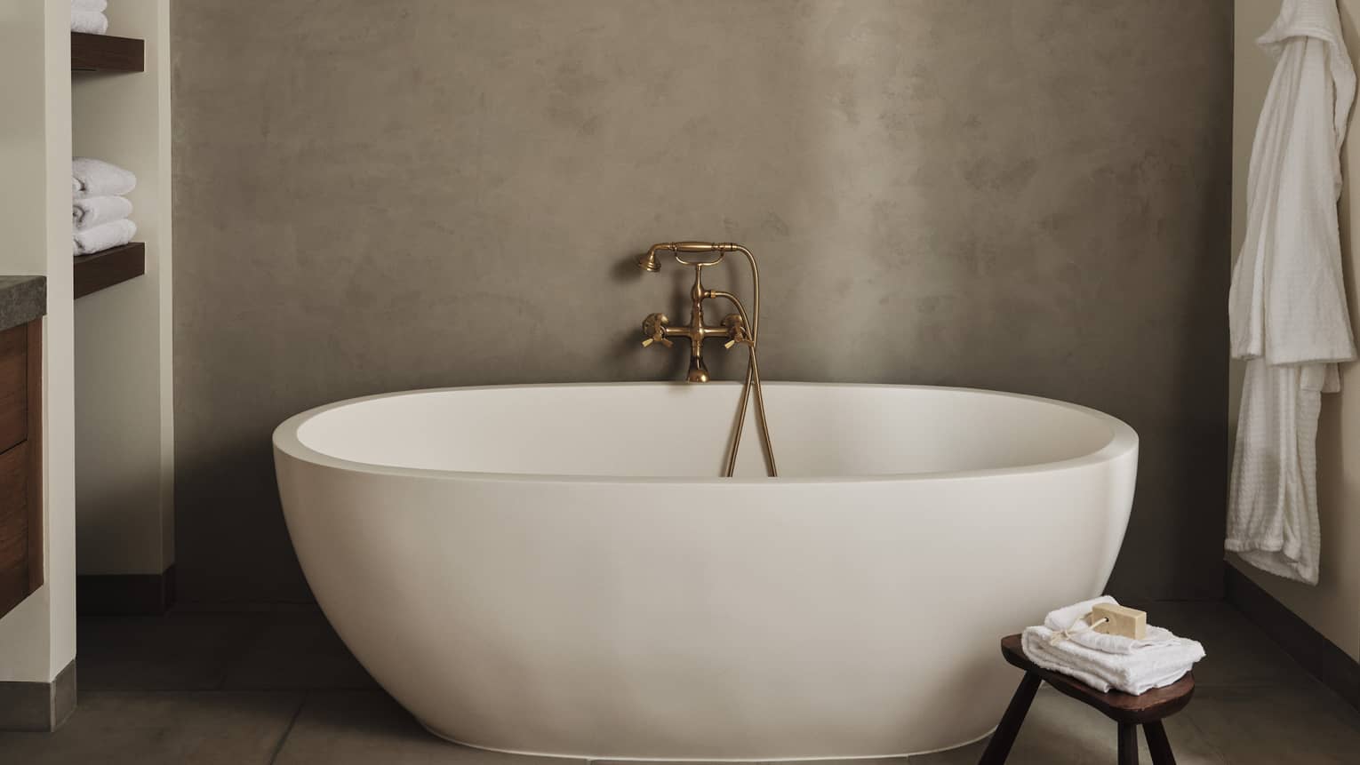 Luxury resort bathroom with a freestanding oval bathtub, built-in towel shelves, small wooden stool with folded towels and soap and bathrobe hanging on the wall