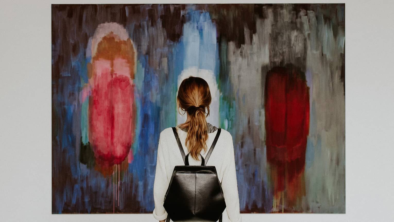 A person with a backpack standing in front of an abstract painting in an art gallery.
