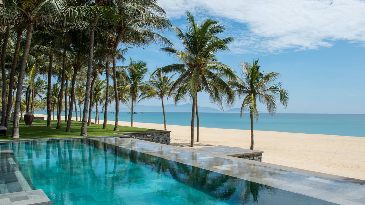 Tall palm trees between stone outdoor swimming pool and white sand beach