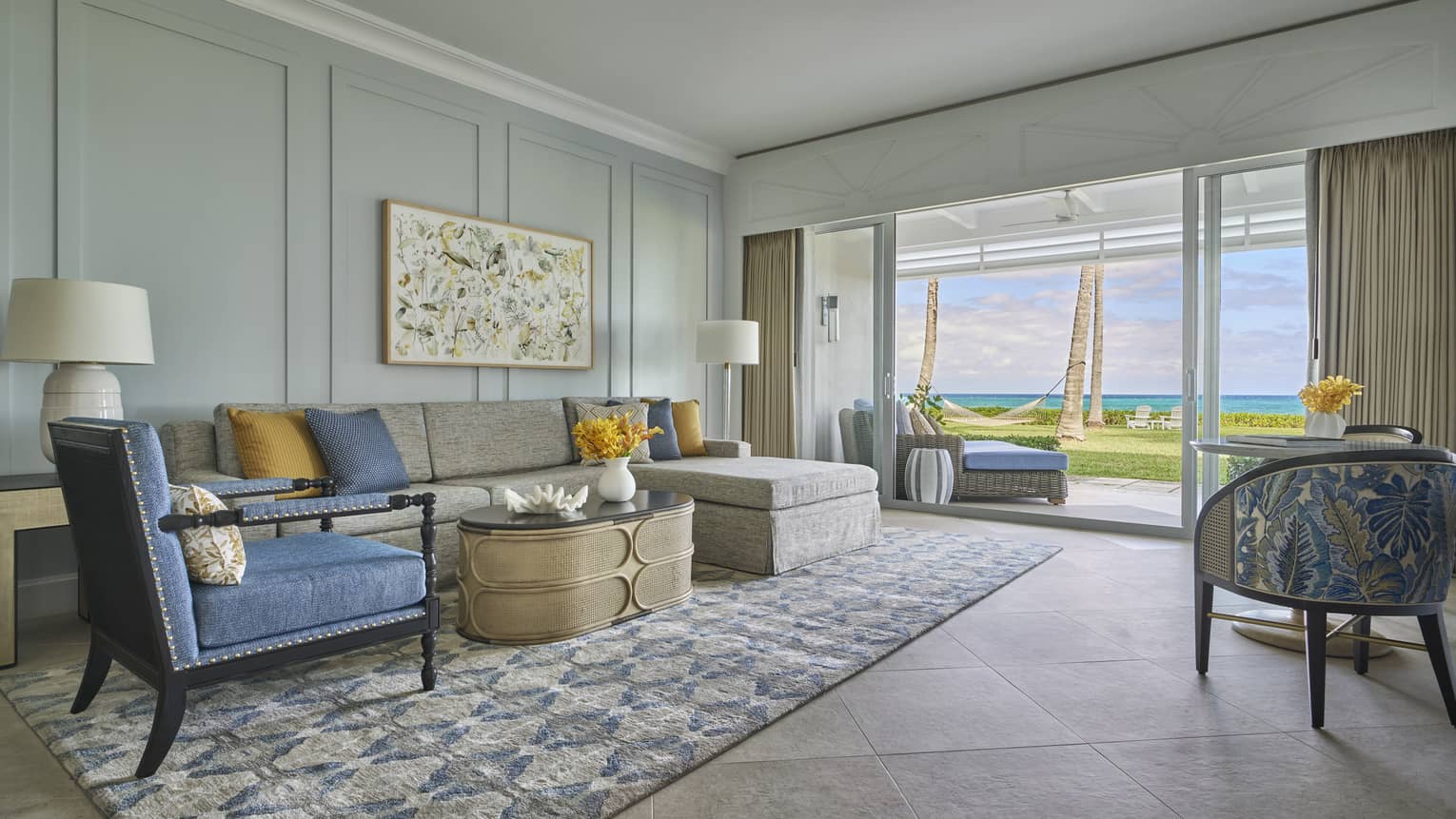 Living room with a gray sectional sofa, blue armchair, patterned rug and glass doors opening to an ocean view with palm trees