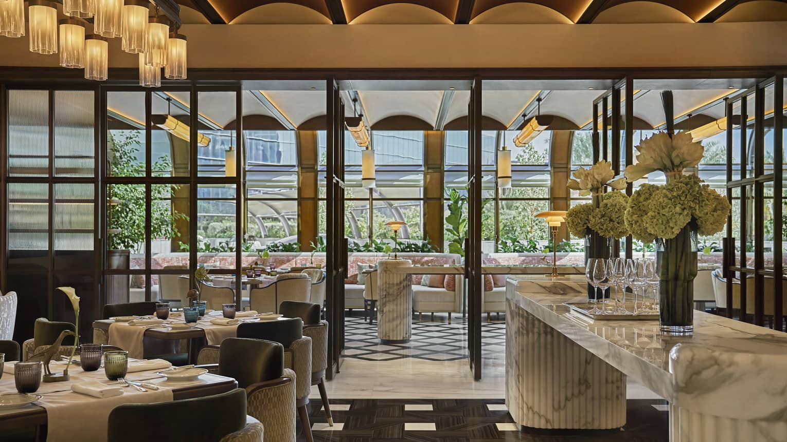 Sun-lit dining restaurant dining room with black-and-white tile flooring, a large white marble counter and floor-to-ceiling glass doors that lead out to a terrace with more seating