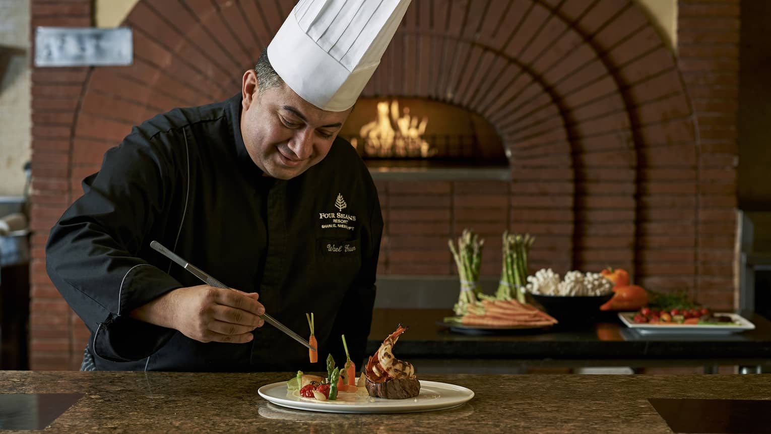 Chef in uniform holds tweezers, arranges vegetables on plate with large steak, lobster tail by wood-burning oven
