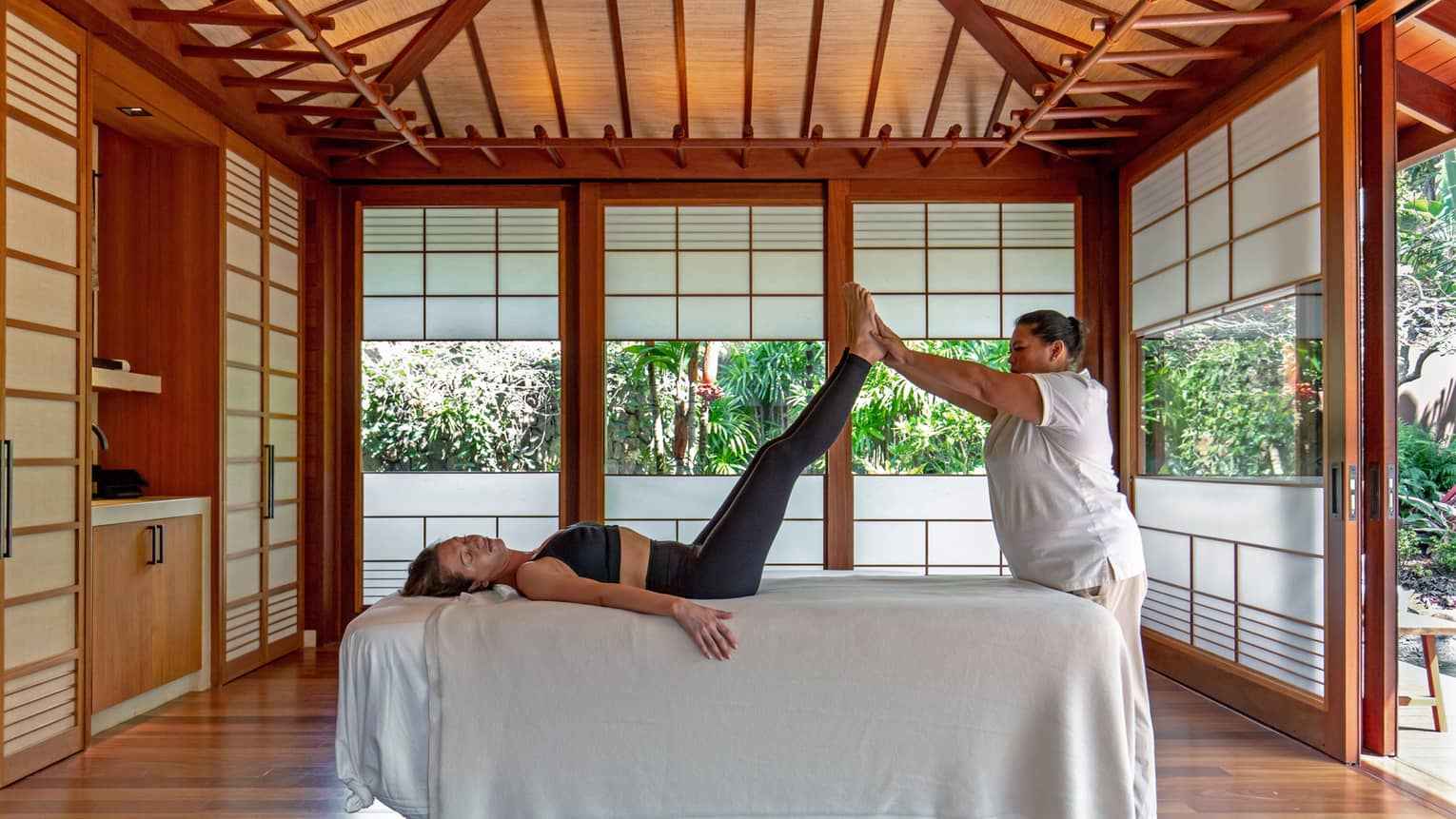 Wellness practitioner lifts legs of woman on spa treatment bed, in a private hale with tropical surroundings