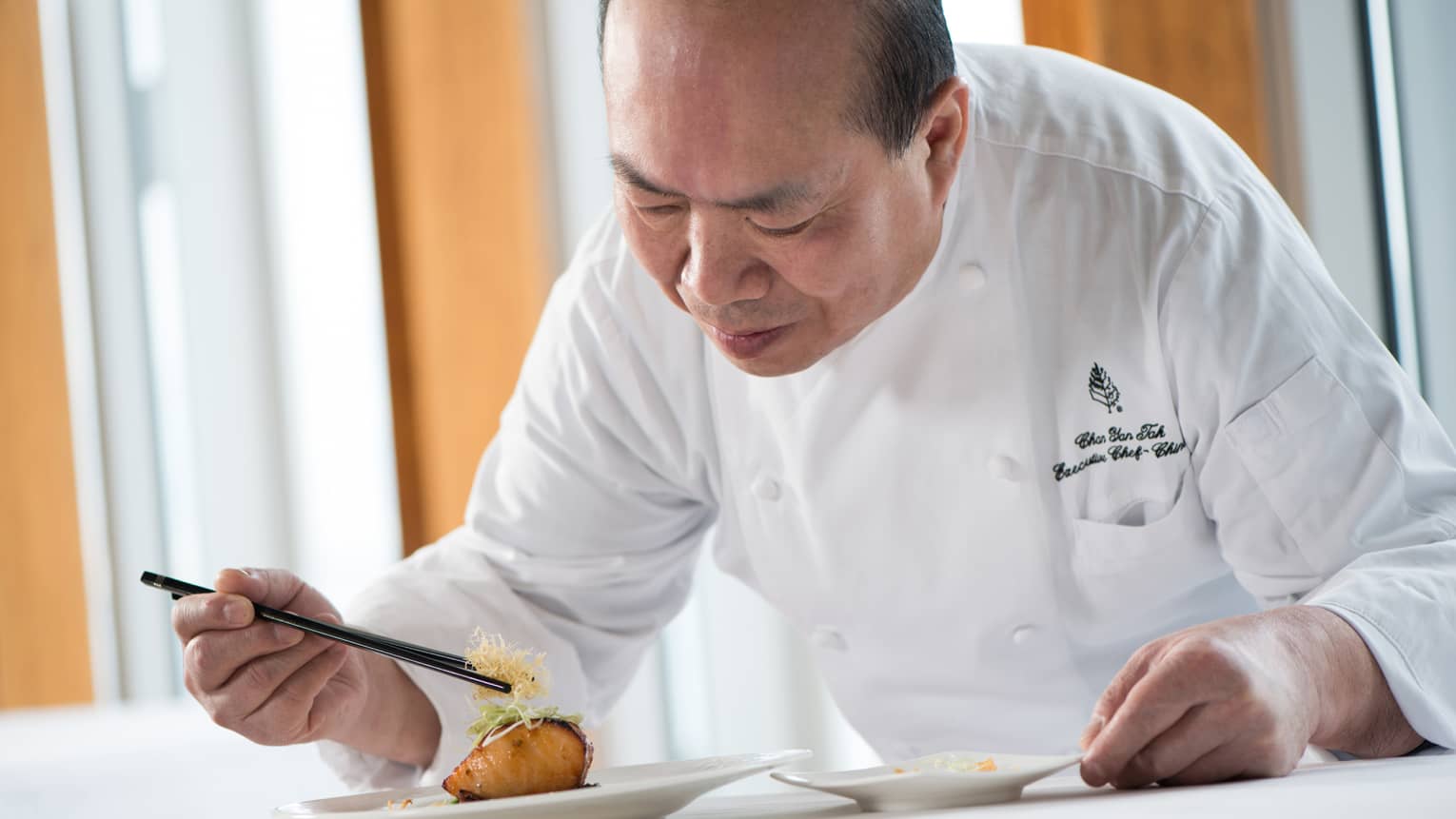 Chef in white uniform leans over grilled salmon dish on plate, adds garnish with chopsticks