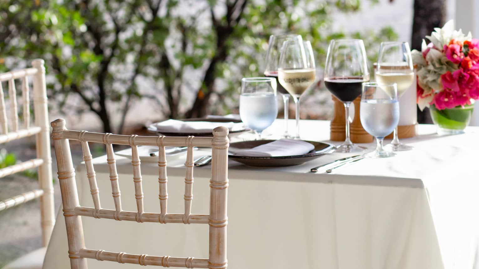 Table for two set with white linens, glasses of wine, and flowers; a low tree and a beach sit just out of focus beyond.