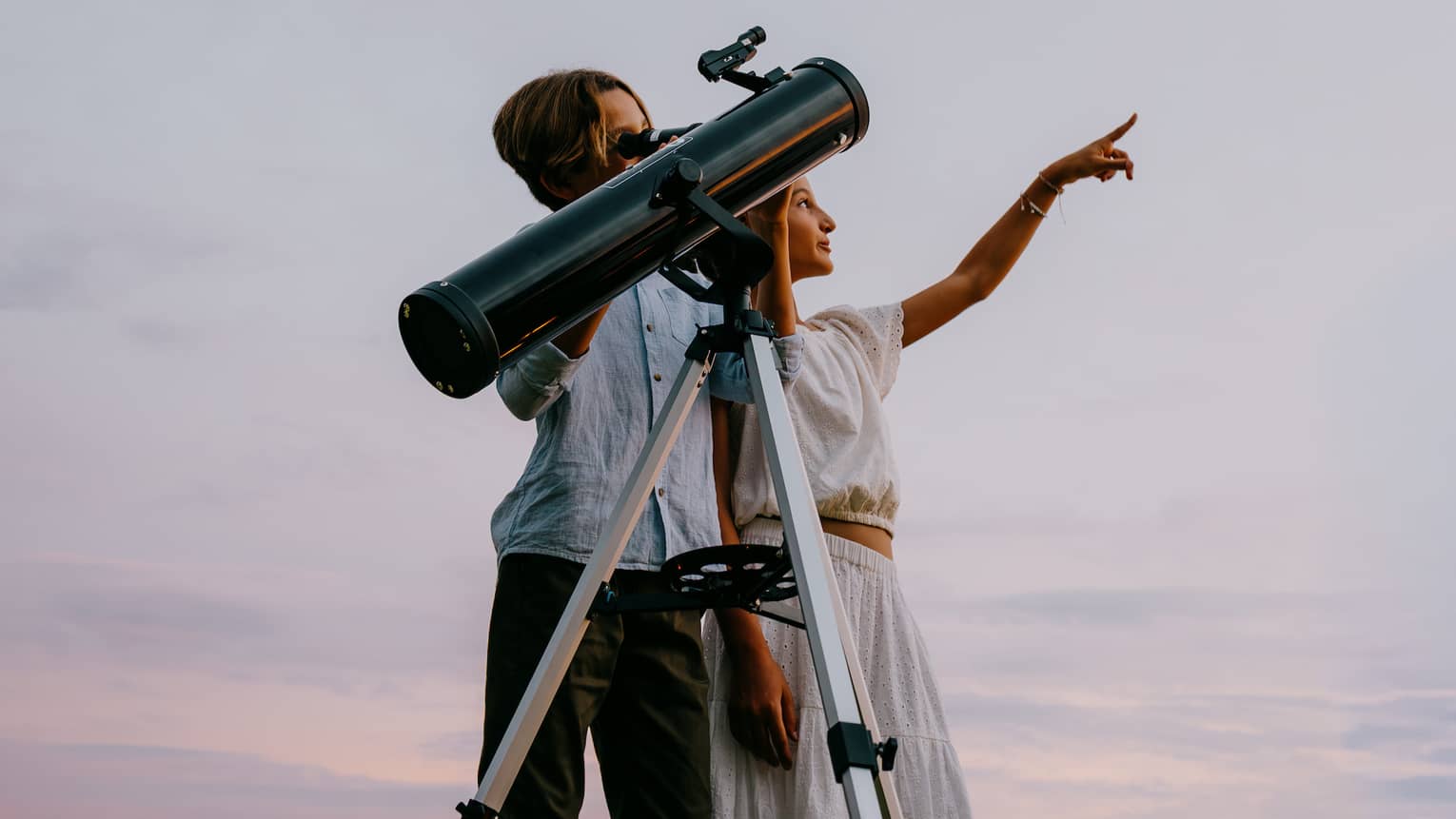 Two people stand next to a large telescope with an evening sky in the background. One person points toward the sky.