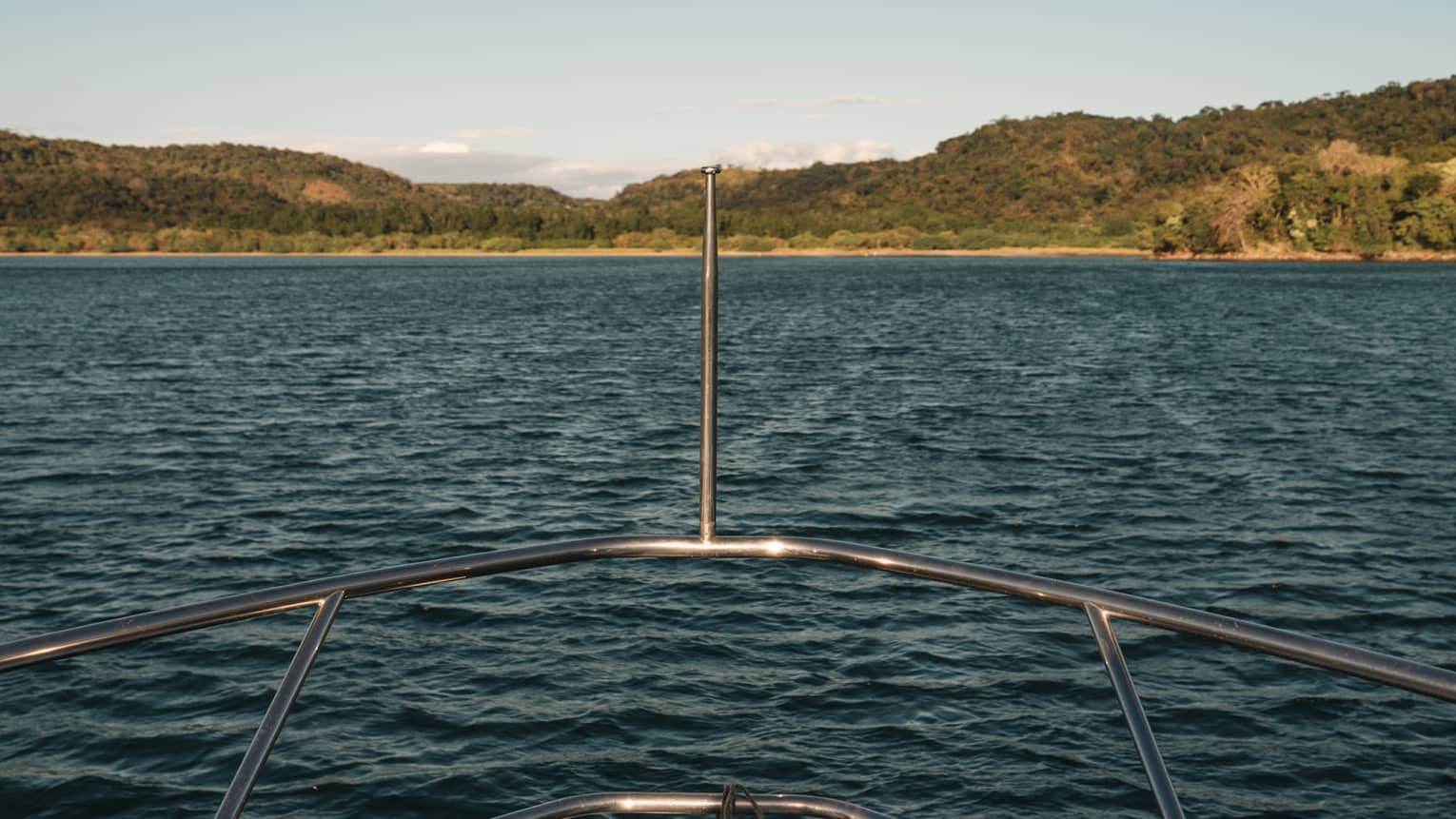 View from the stern of a boat, where overhead a sea bird floats above rippling water towards a distant forested island.