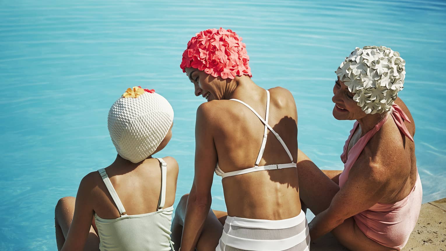 Three people in swim caps sitting on the edge of the pool.