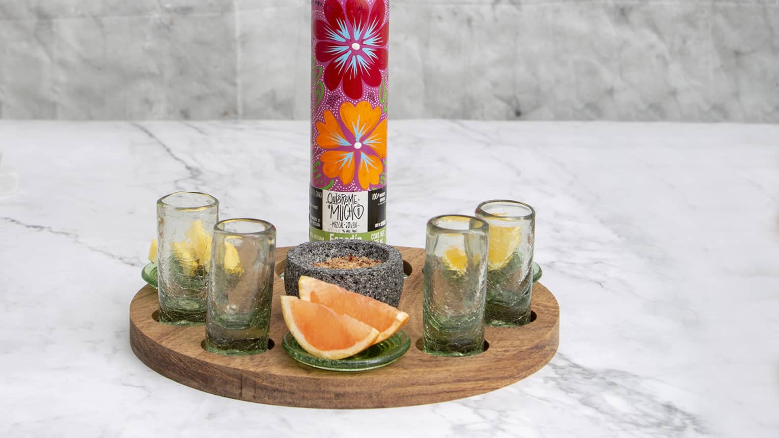 Brightly floral-painted mezcal bottle on wooden tray with four shotglasses, stone spice bowl and lemon and grapefruit wedges.