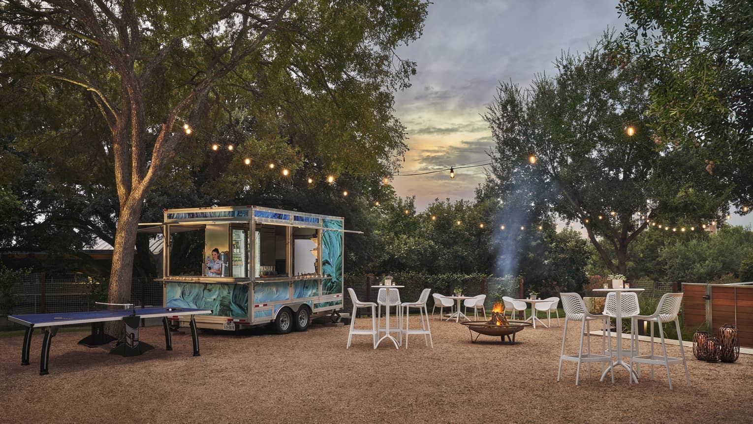 A food truck outsider surrounded by chairs, tables and trees.