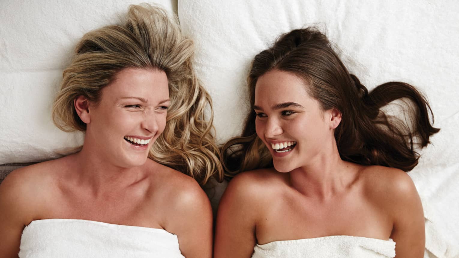 Two laughing women wearing white towels lie on massage table in spa