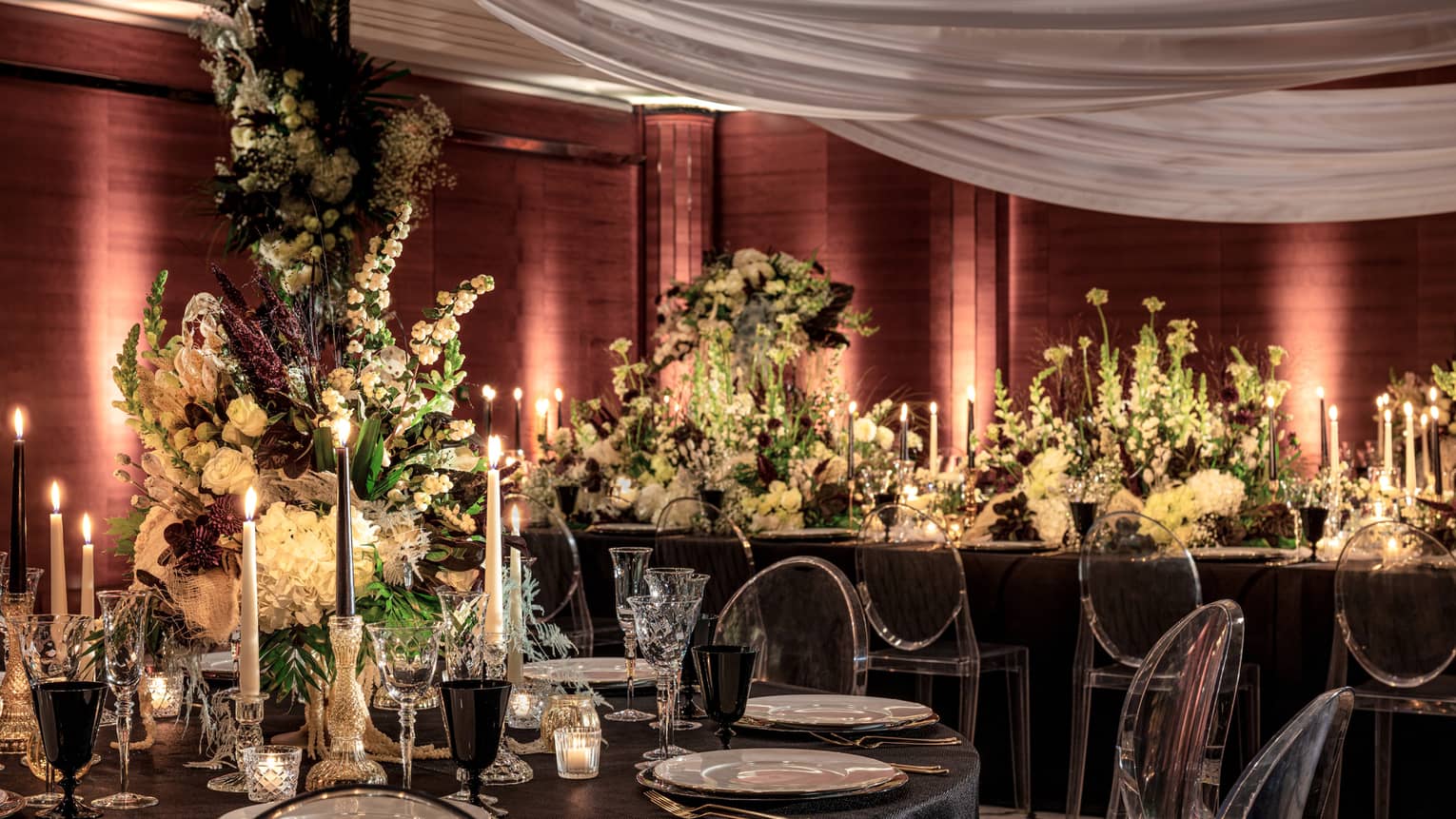 A sumptuously decorated ballroom at the Four Seasons London Park Lane in Mayfair, set for a wedding reception. The venue features dark, moody lighting complemented by rich, deep red drapery that envelops the room. Each table is elegantly set with black linens, ornate floral centerpieces of yellow and white flowers, and numerous lit candles, creating a romantic and luxurious atmosphere. Overhead, sheer white fabric swathes across the ceiling, enhancing the sophisticated and intimate ambiance of this enchanting wedding venue.