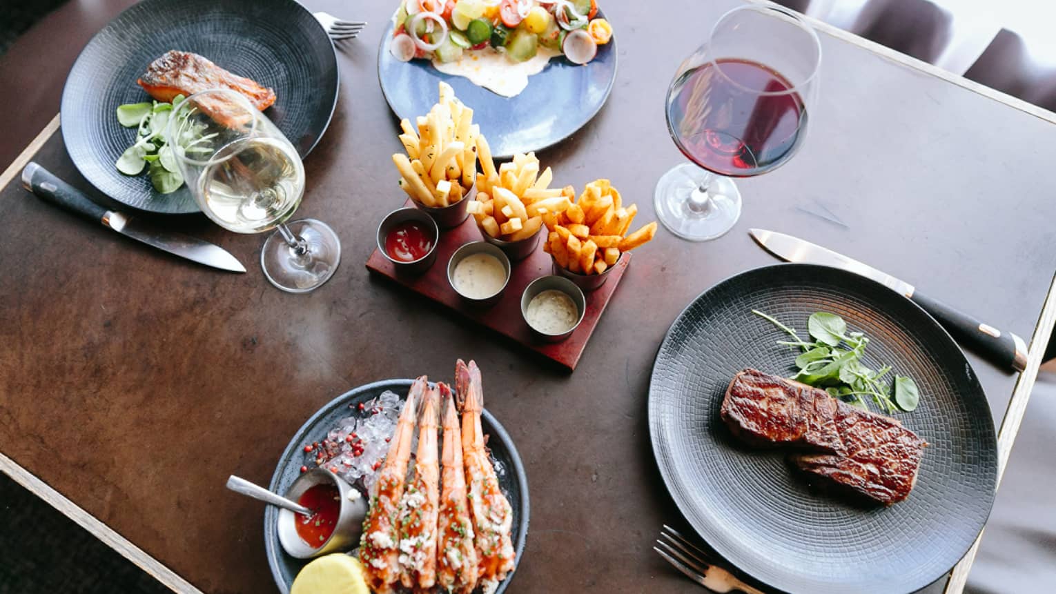 A wood table with four plates of food including steak, seafood, fries, and fish.
