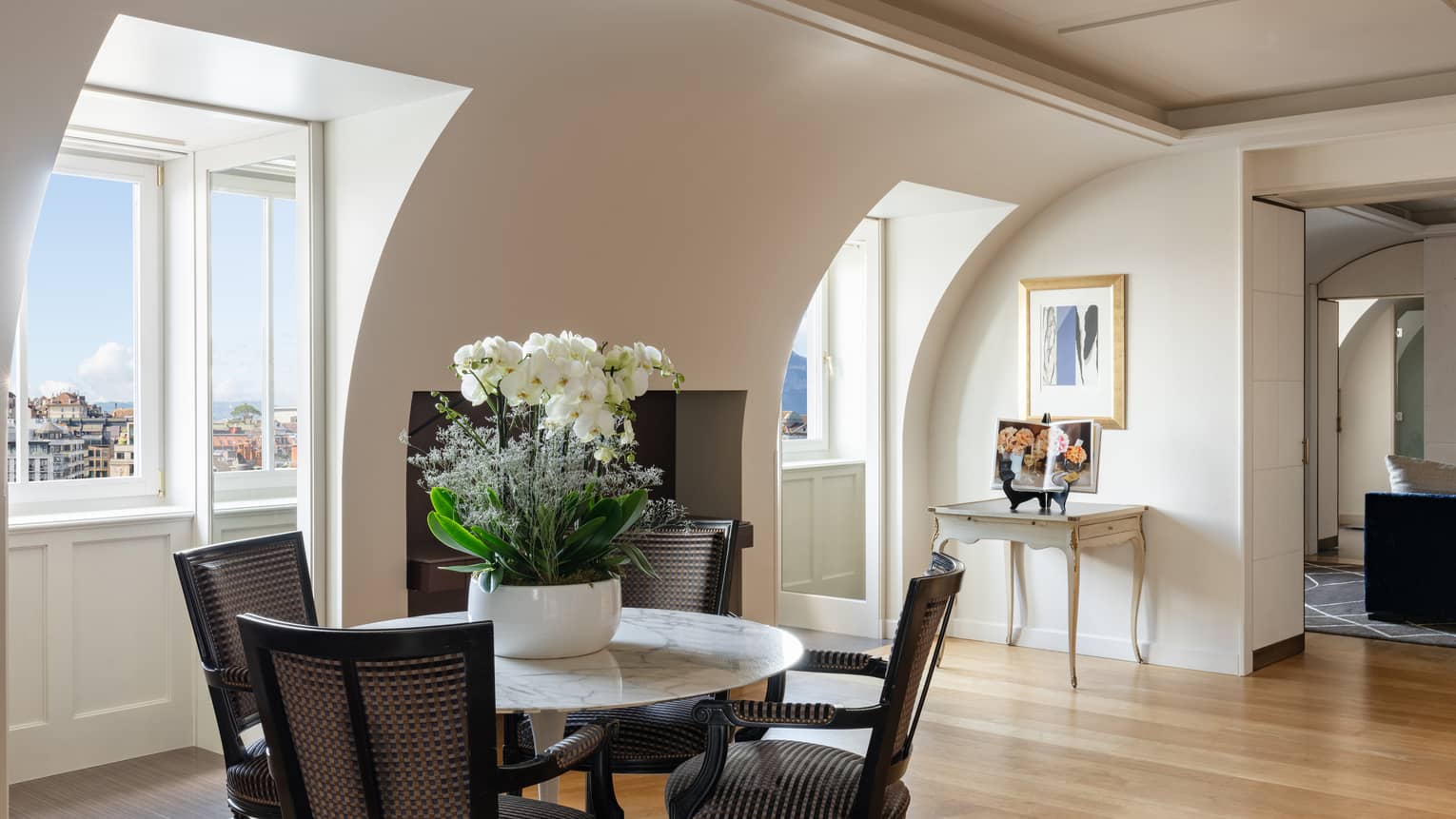 Luxury hotel suite dining area with a round marble table with four chairs and floral arrangement, set near windows with archways offering city views 