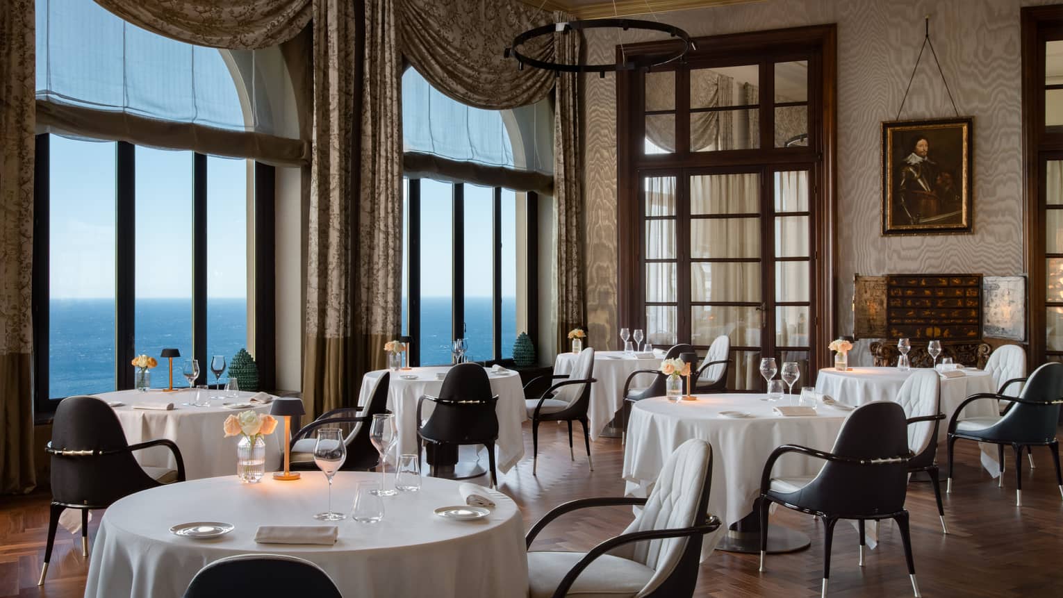 Indoor restaurant with white tablecloths, large drapery framing windows that look out to sea