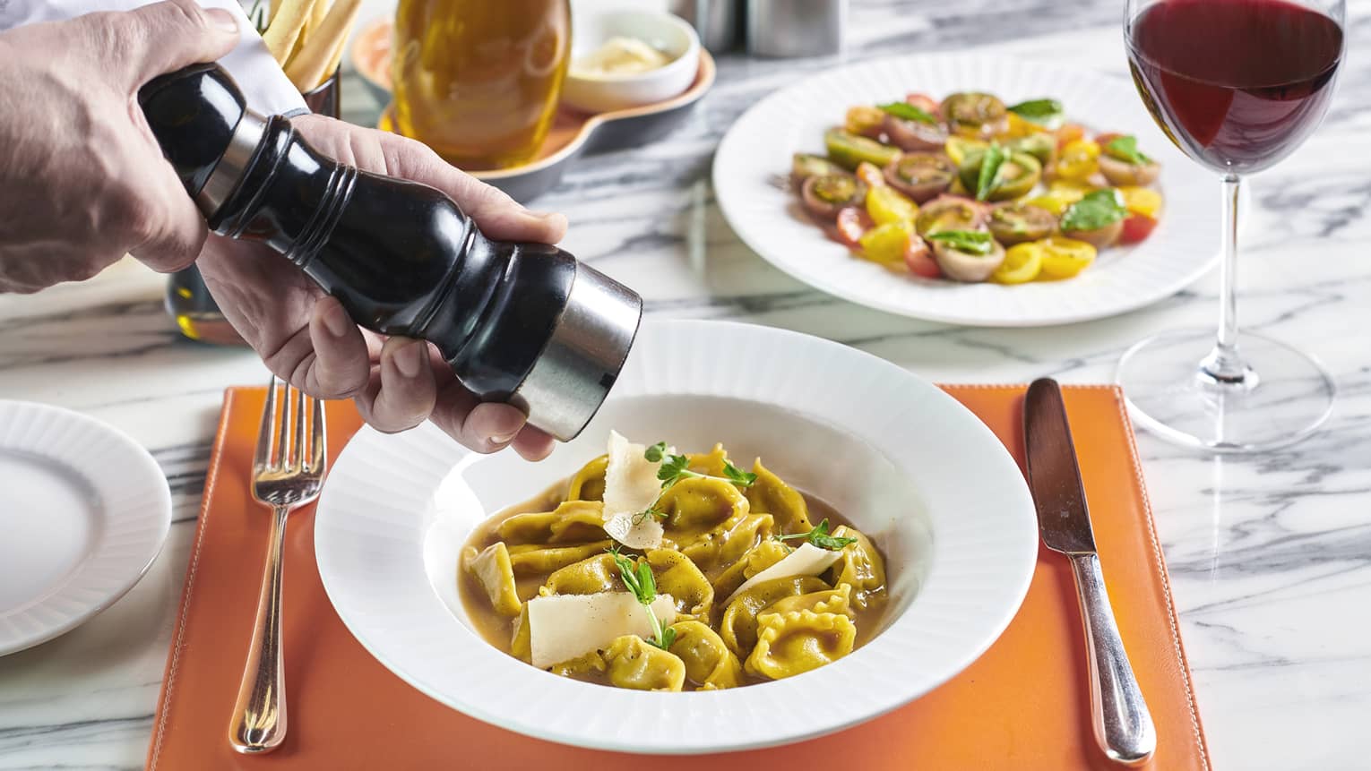 Hands grinding pepper over pasta dishes on white marble table with red wine glass
