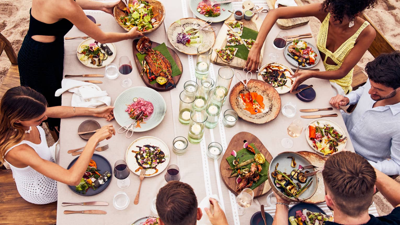 Beachside dining table with an assortment of vibrant dishes, candlelit glass holders and guests enjoying fresh seafood and grilled vegetables