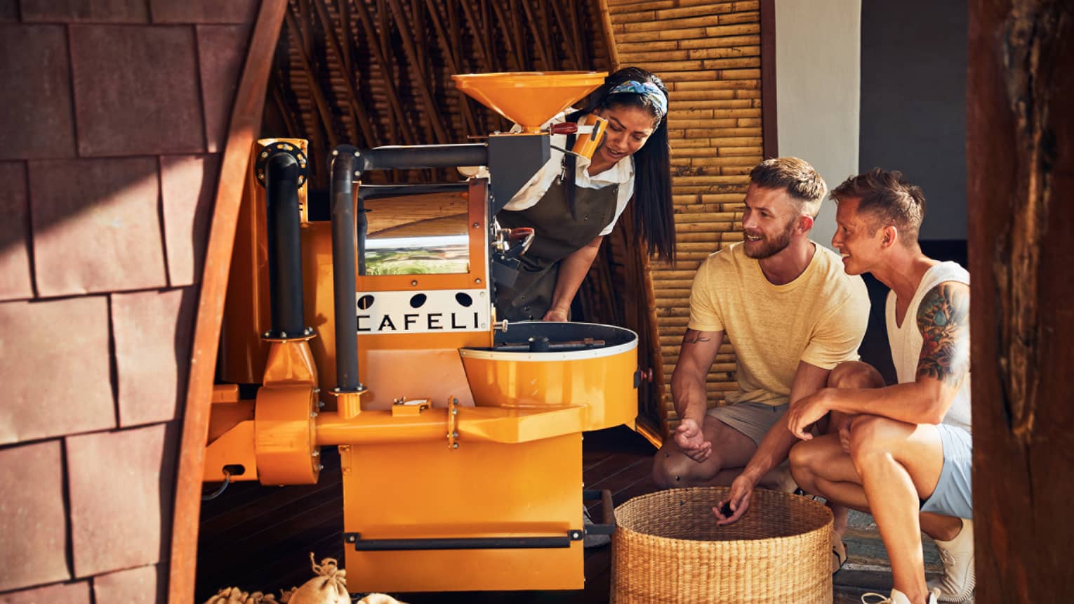 A woman demonstrates coffee roasting to two guests, who observe closely near a yellow coffee roaster in a bamboo-walled setting.
