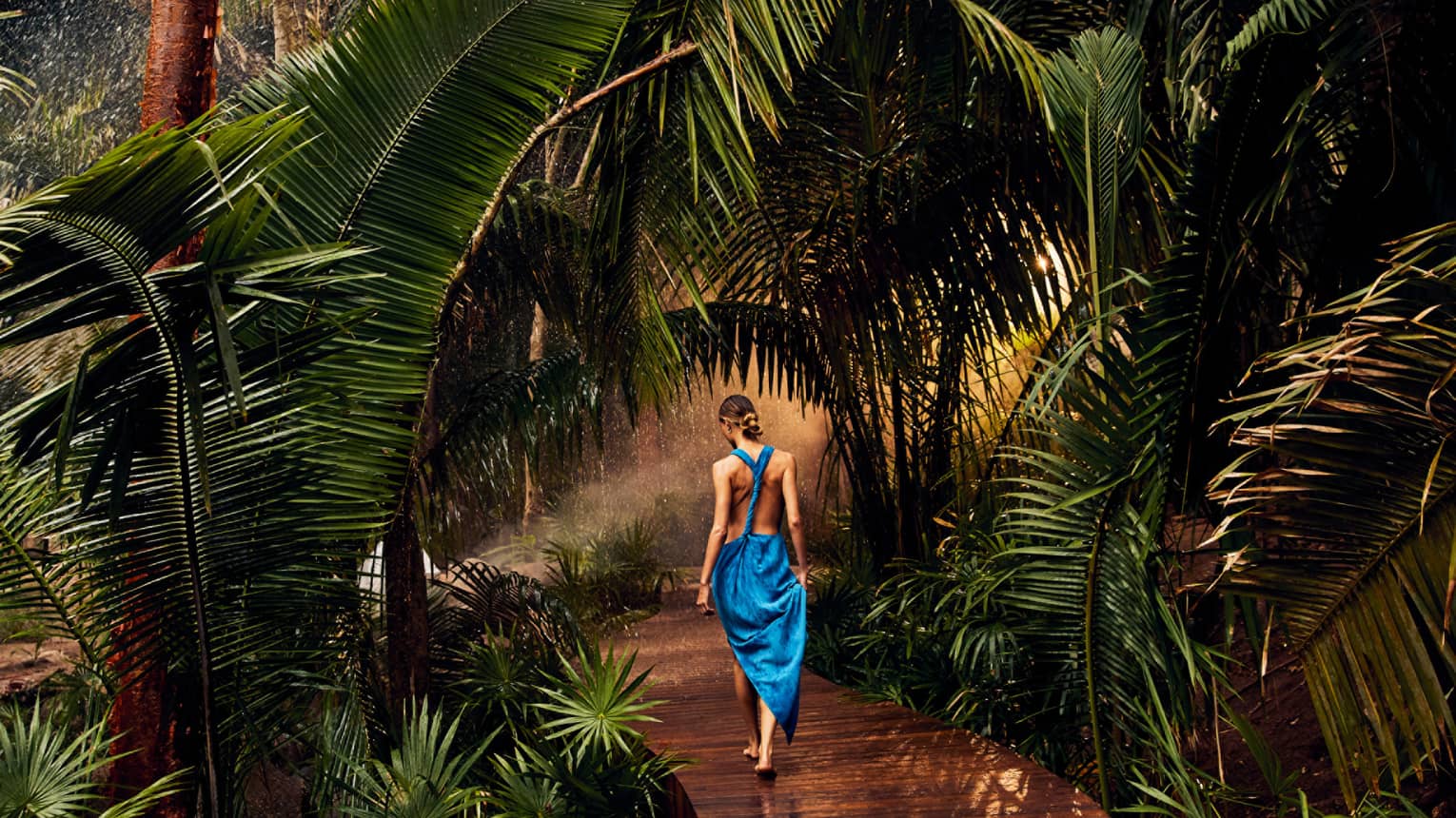A woman in a blue dress walking along a wooden path covered in palm trees.
