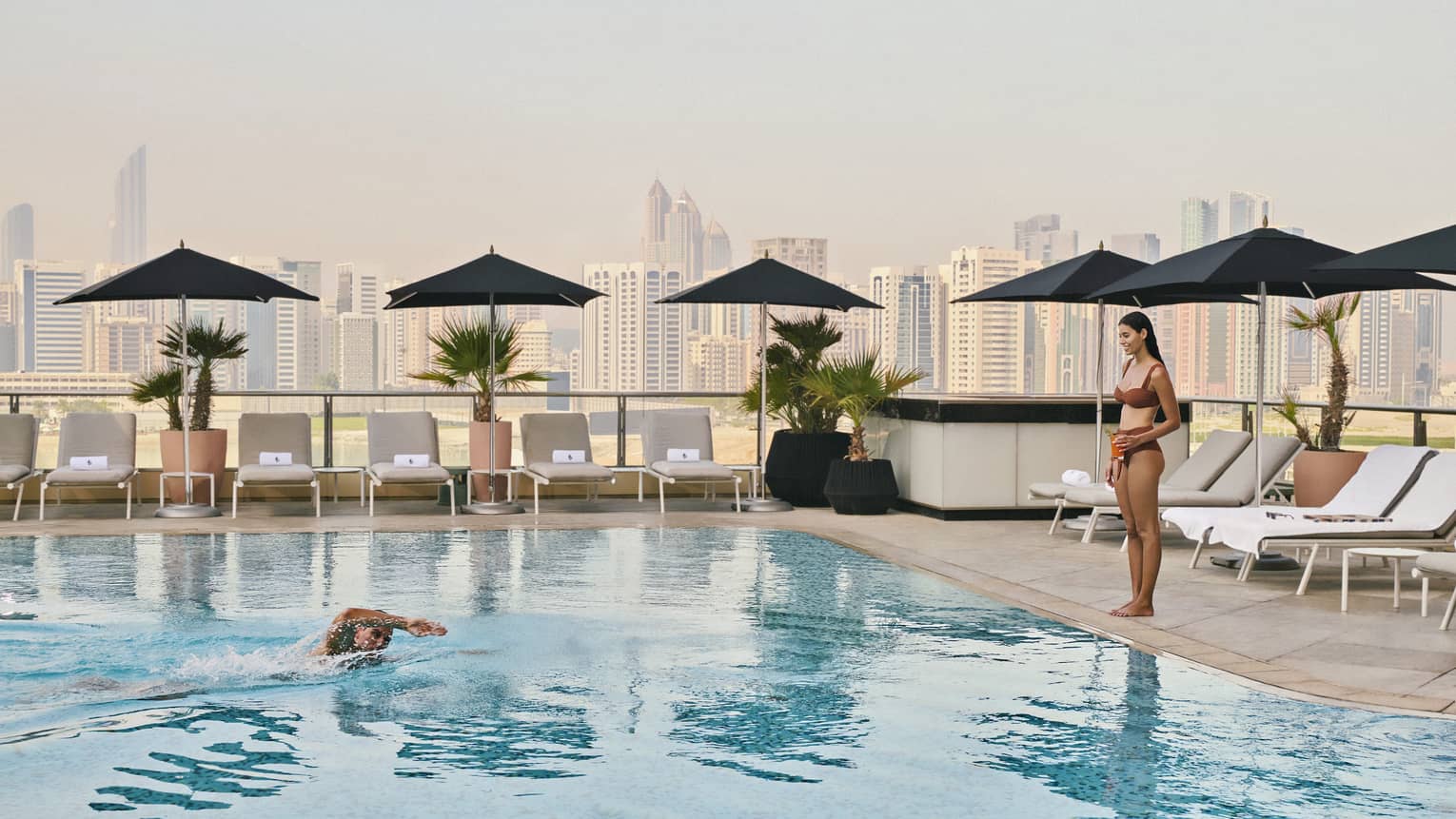 One guest stands by a rooftop pool while another swims in the water