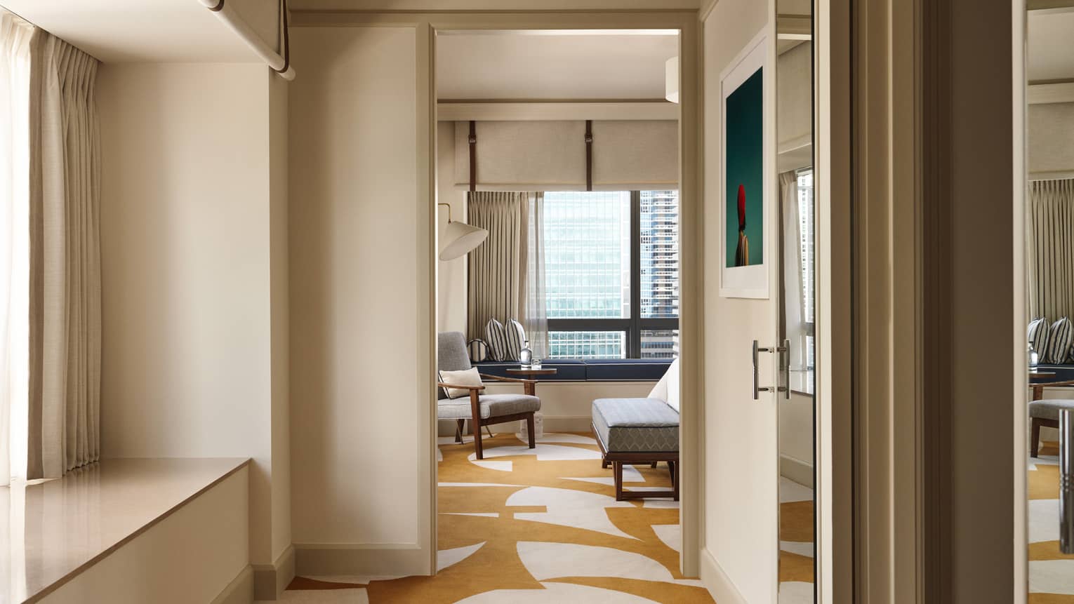View through an open doorway into a luxurious one-bedroom suite at the Four Seasons Hotel in Miami, showcasing a modern living area with stylish furniture, including a plush grey sofa and matching armchairs. The suite features a geometric patterned rug and large windows that provide a sweeping view of the city skyline. The interior design combines contemporary elegance with comfort, highlighted by soft, natural lighting and neutral colour tones.